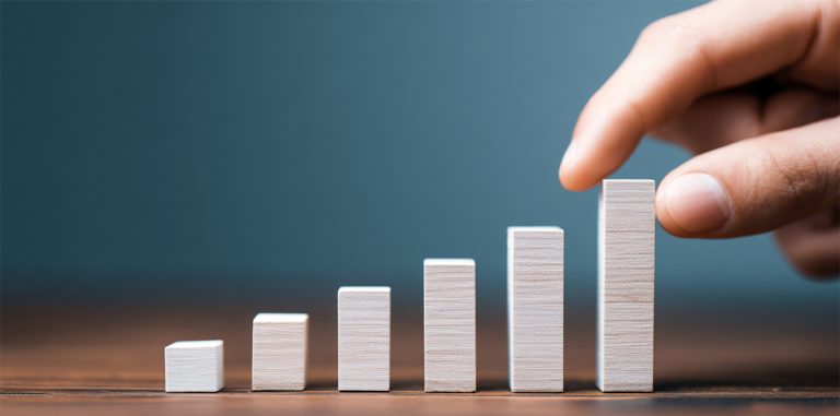 Hand placing wooden blocks in ascending order from left to right, creating a bar chart pattern that represents incremental growth and improvement against a teal background.